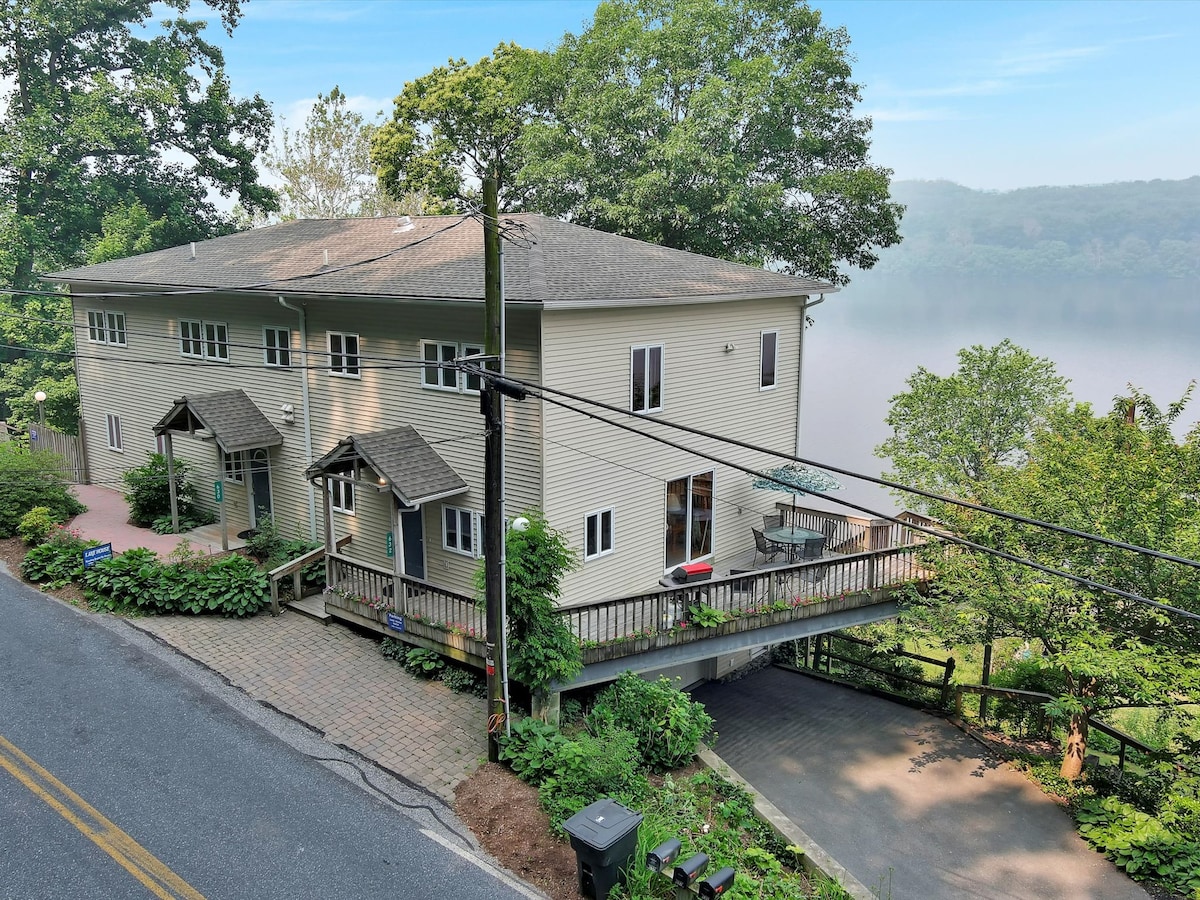 A lakefront townhouse is visible, featuring multiple entrances with overhangs and a wraparound deck. Lush greenery surrounds the property, complementing the serene view of the lake in the background. Paved walkways lead to the entrances, enhancing accessibility in this tranquil nature setting.