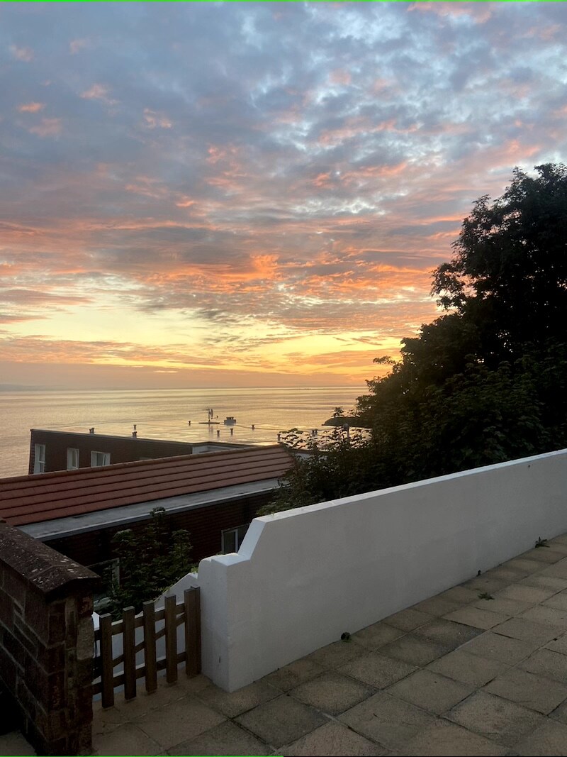 A sunset view over the sea is captured from a terrace. Soft orange and pink hues fill the sky, reflecting on the water. Lush greenery frames the edge of the terrace, while a low white wall provides a barrier along the patio.