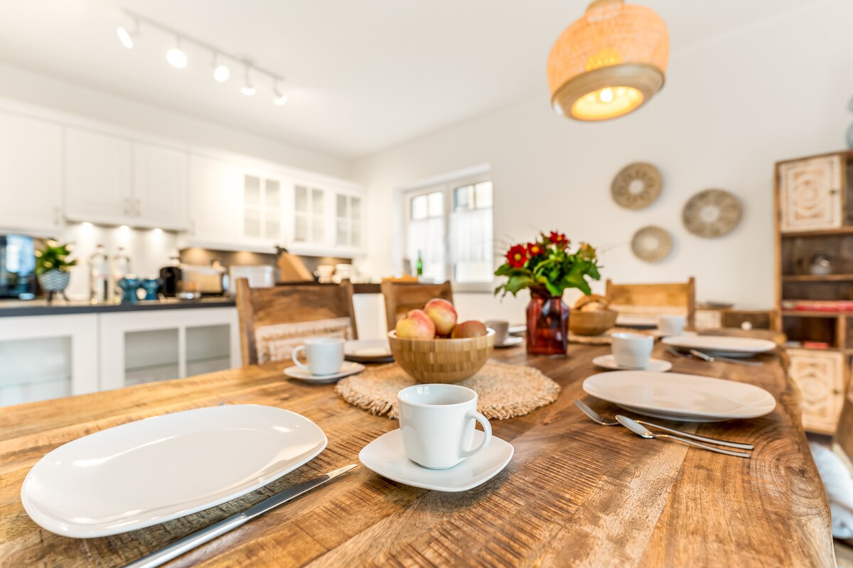 A rustic wooden dining table is set with white plates and cups, complemented by a central bowl of apples. The kitchen in the background showcases modern appliances and ample storage. Natural light illuminates the space, enhancing the inviting atmosphere with decorative wall accents.