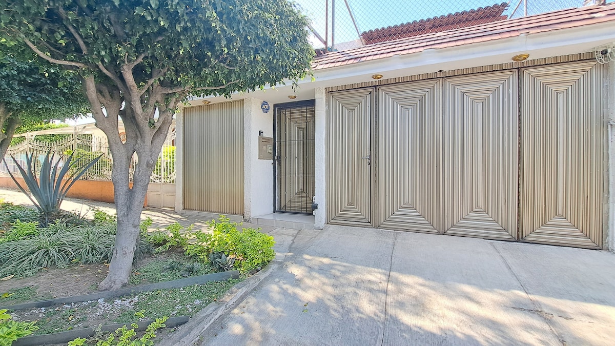 The exterior view of the property showcases a well-maintained entrance with golden sliding gates. Lush greenery and small shrubs line the pathway, leading to the main door. A tree provides shade on one side, enhancing the inviting ambiance of the entrance.
