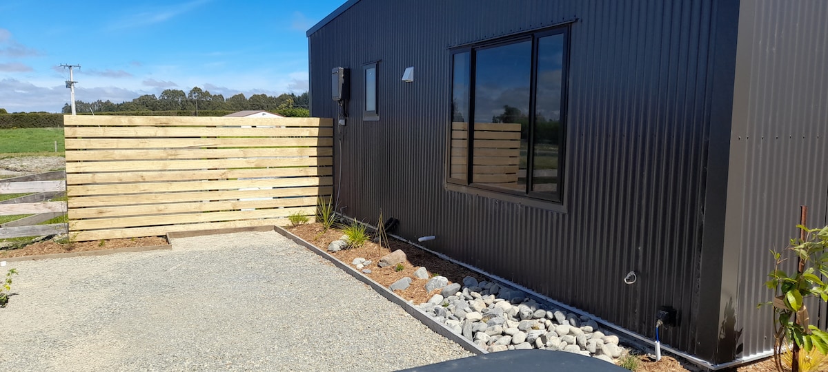 A modern, black exterior structure is depicted alongside a gravel pathway. A wooden fence provides privacy, while neatly arranged stones and plants enhance the outdoor area. A clear blue sky frames the scene, contributing to a peaceful rural atmosphere.