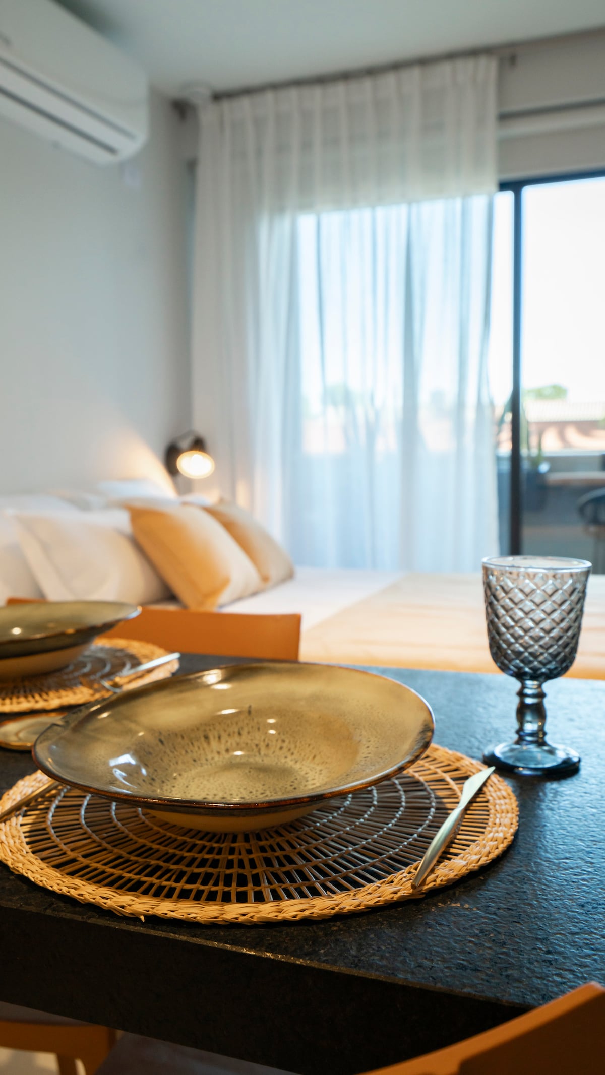 A dining table is set with a textured bowl and a clear glass goblet. Soft light filters through sheer curtains, illuminating the cozy sleeping area in the background, where a spacious bed is dressed in neutral tones.