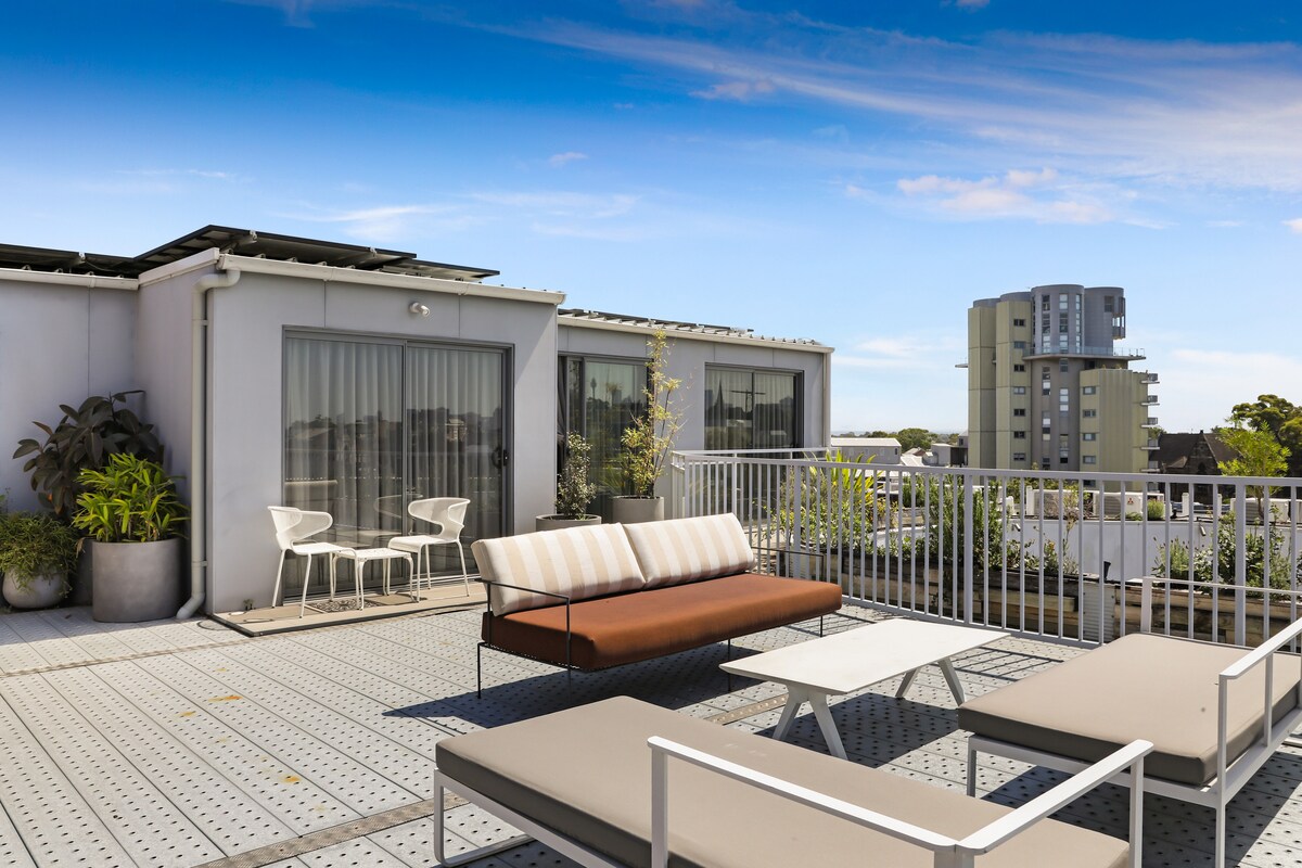 A spacious rooftop terrace is presented, featuring a comfortable lounge area with a stylish sofa and sun loungers. Potted plants add greenery, while a pair of chairs and a small table invite relaxation. Panoramic views of neighboring buildings and the sky are visible.