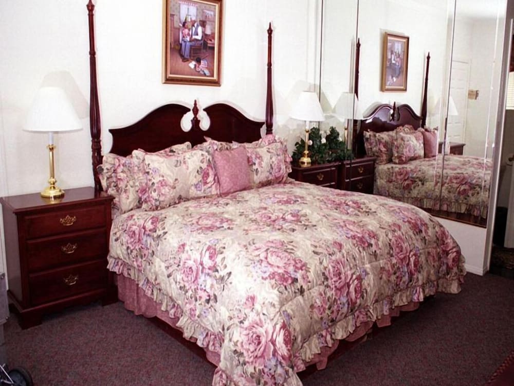 A well-furnished bedroom features a spacious bed adorned with floral-patterned bedding and decorative pillows. A mirrored wall enhances the room's brightness, while a bedside table with a lamp and a small decorative plant adds functionality.