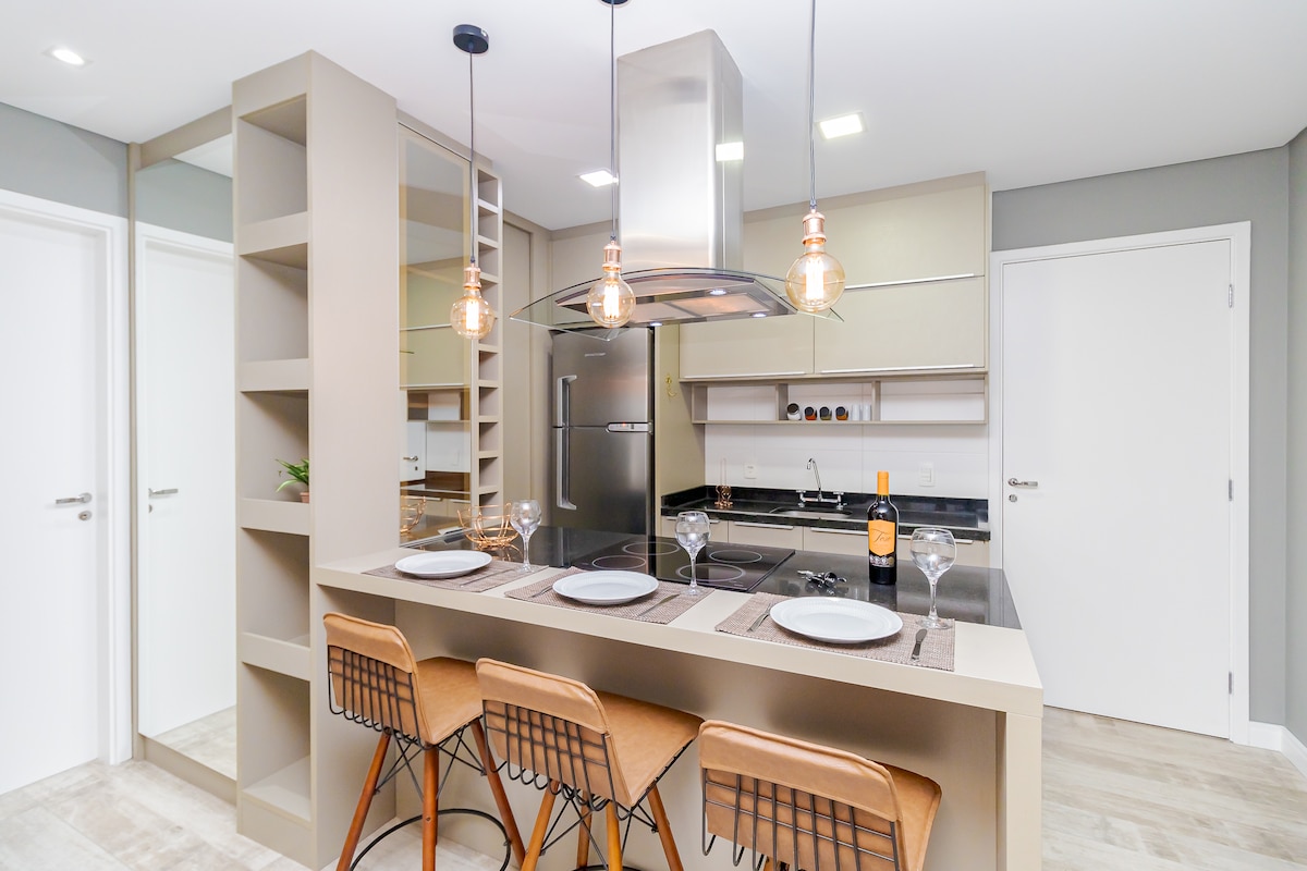 A modern kitchen area is featured with sleek cabinetry and polished countertops. Three bar stools are positioned at the island, complemented by pendant lighting above. A stylish wine bottle is placed on the counter near elegant glassware, enhancing the functional design of the space.