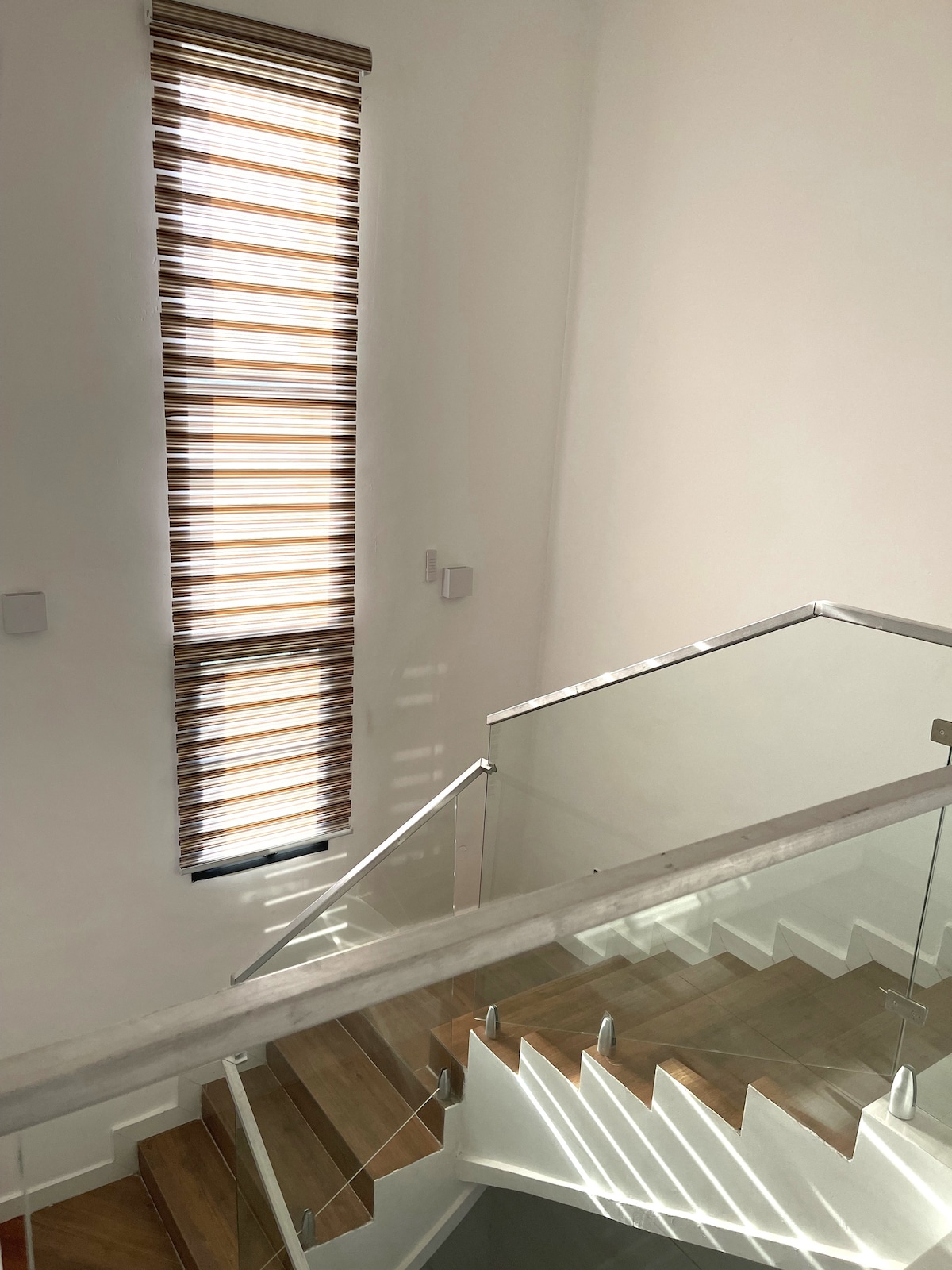 A well-lit staircase is featured with sleek glass railings and wooden steps. Natural light streams through a tall window dressed in horizontal blinds, casting soft shadows on the walls. The minimalist design enhances the spacious feel of the area.