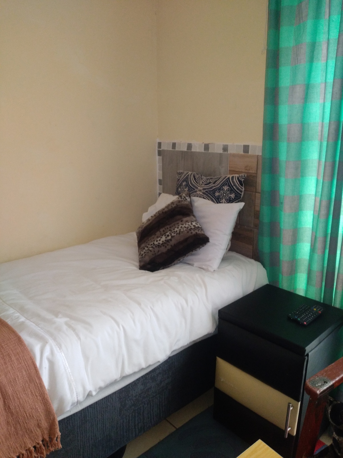 A cozy sleeping area features a single bed with a white coverlet and decorative pillows. A green and gray checkered curtain filters light from a window, while a bedside table with a remote control adds functionality to the space.
