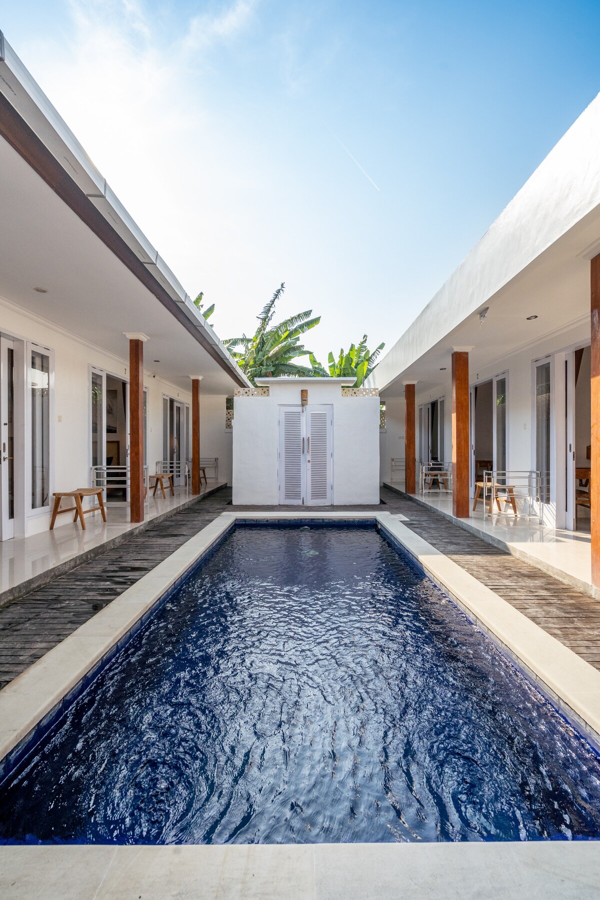 A serene outdoor pool is framed by two guesthouse structures, featuring modern architecture with large glass doors. Lush greenery is visible in the background, and seating areas are positioned along the poolside, inviting relaxation and enjoyment in this tranquil setting.