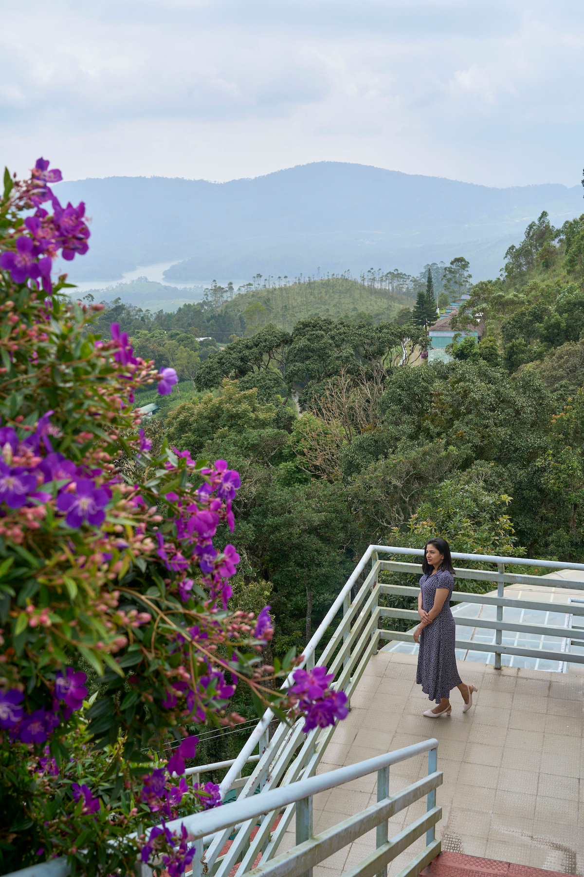 A balcony offers a scenic view of rolling hills and lush greenery, framed by vibrant purple flowers. A pathway leads off the balcony, enhancing the sense of openness and connection with nature.