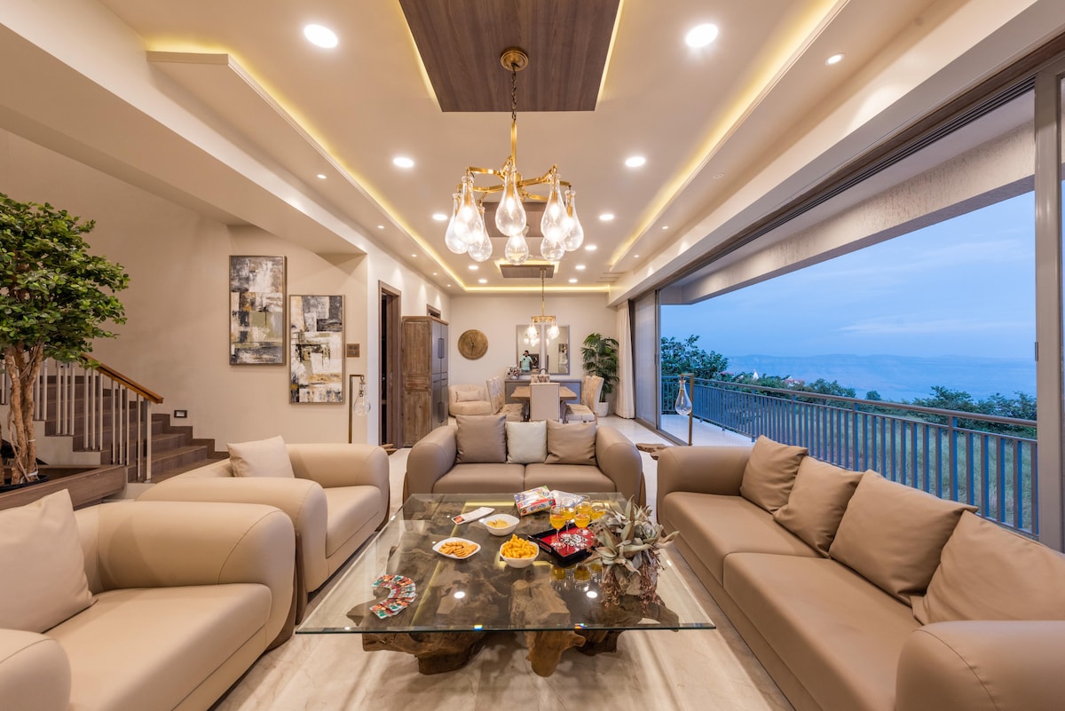 A spacious living area features plush taupe sofas arranged around a wooden coffee table. Floor-to-ceiling glass doors offer a view of the balcony and the valley beyond. Soft lighting and artistic decor create a bright and welcoming ambience.