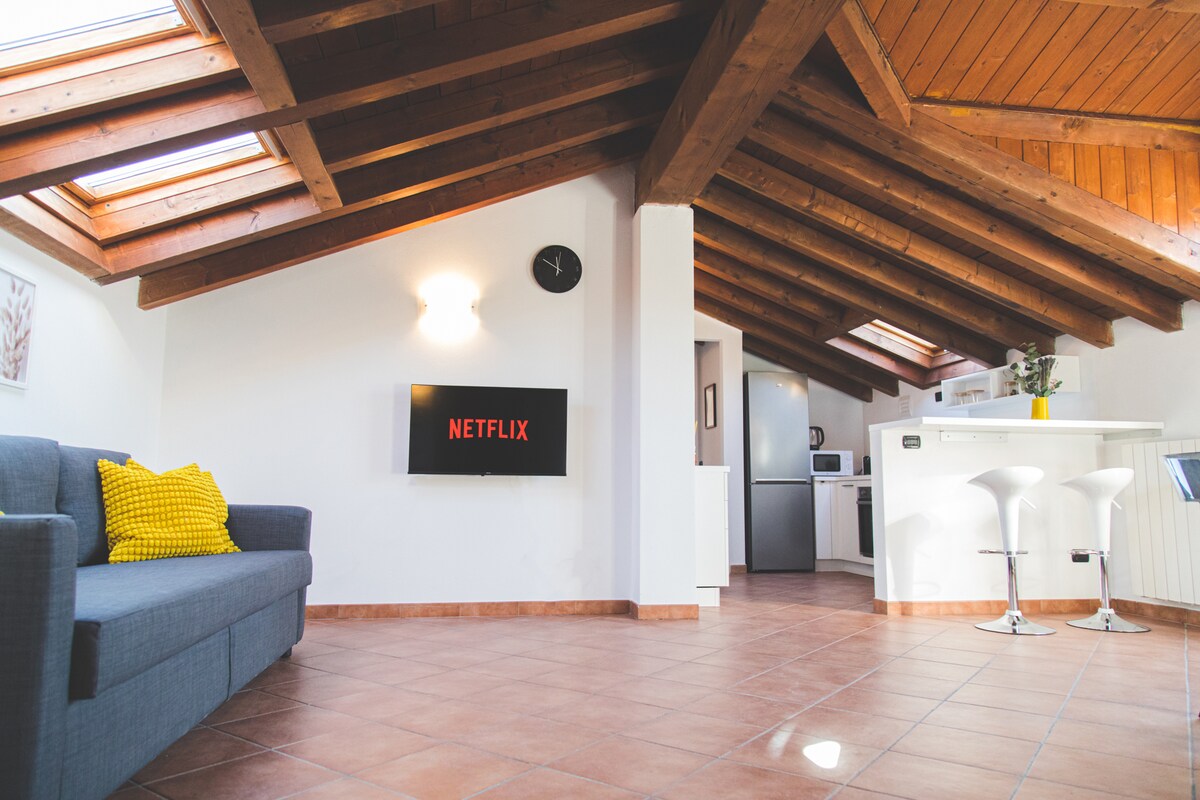A spacious living area is illuminated by natural light from skylights above. A dark gray sofa with a yellow accent pillow is positioned in front of a wall-mounted television displaying the Netflix logo. A modern kitchen bar with two stools is visible in the background.