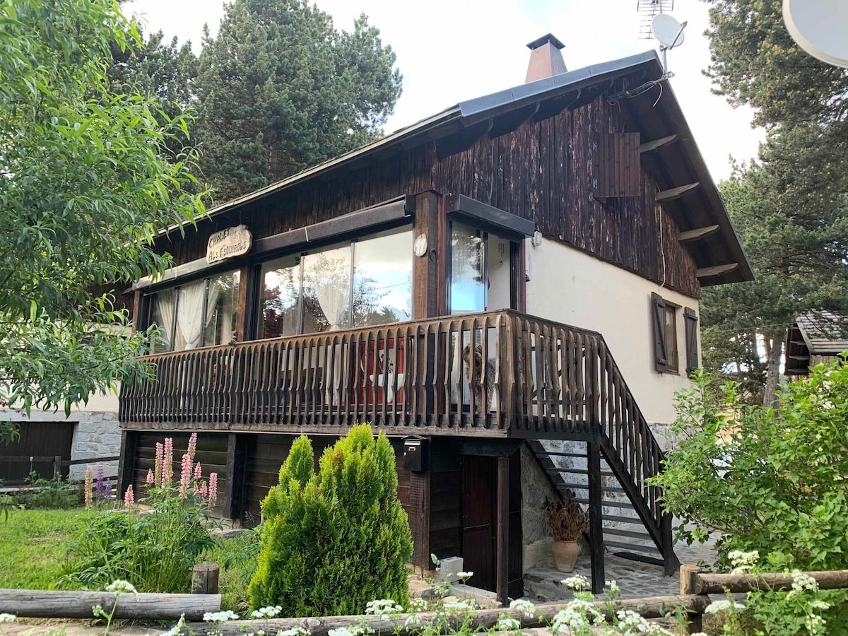 A charming chalet features a covered deck with wooden railings, surrounded by a garden with lush greenery and colorful flowers. Large windows provide natural light to the interior, while the stone pathway leads to the entrance, blending seamlessly with the serene mountain environment.