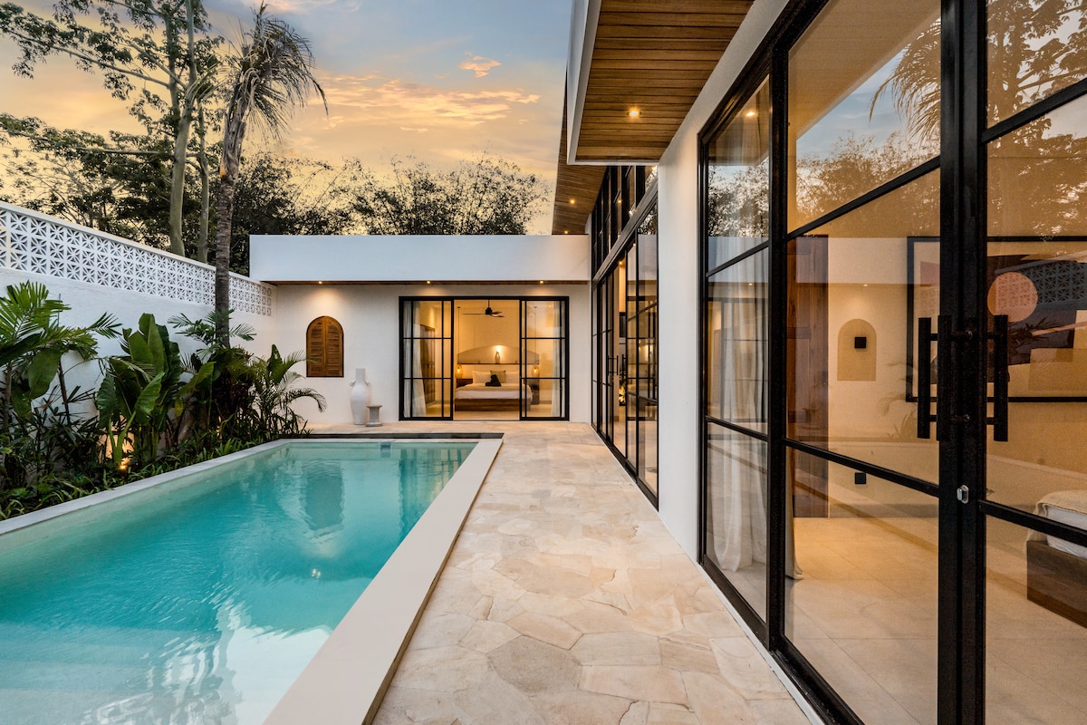 The villa's outdoor space features a swimming pool surrounded by lush tropical greenery. Large black-framed sliding glass doors provide a view of the open living area and tranquil environment. Warm golden light from the exterior highlights the smooth stone pathway alongside the pool.
