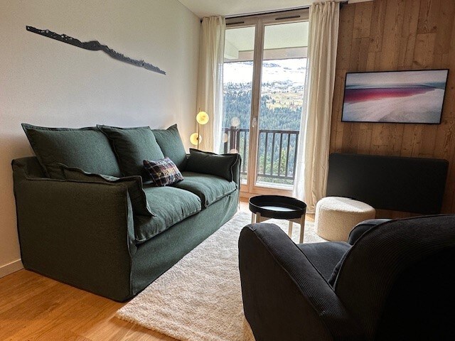 A comfortable living area is presented, featuring a plush green sofa adorned with decorative cushions. A cozy black armchair is positioned nearby. Large windows allow natural light to fill the space, offering a scenic view of the surrounding mountains. A wall-mounted TV is also visible.