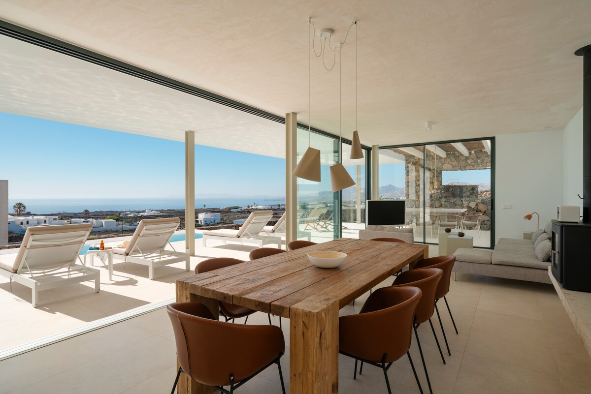A spacious dining area showcases a large wooden table surrounded by brown chairs. Floor-to-ceiling glass doors open to a terrace with sun loungers, offering views of the landscape and sea beyond. Natural light floods the room, creating an inviting space for meals.
