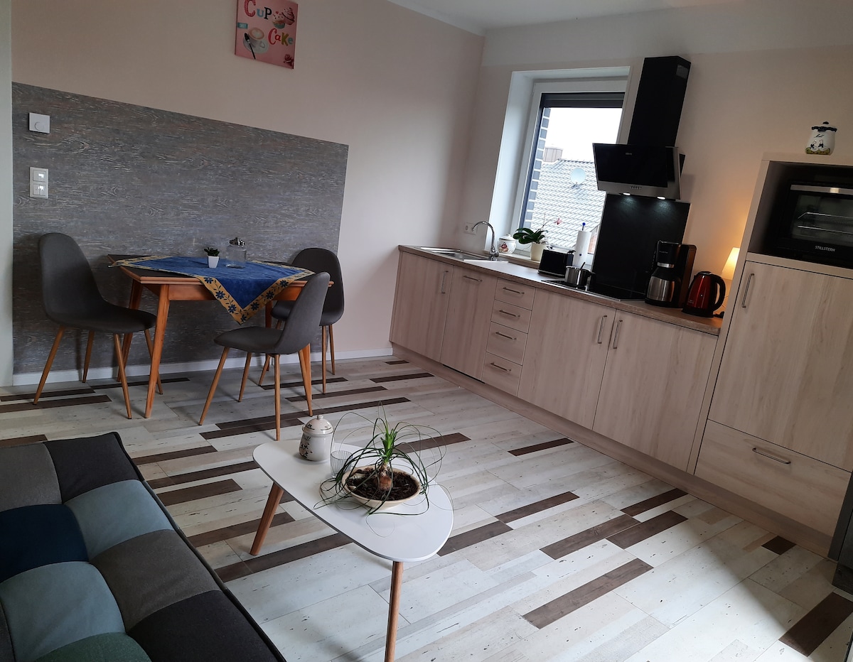A modern kitchen area features light-colored cabinets and a dark countertop, complemented by a sleek black stove and appliances. A small dining table with two chairs sits nearby, accompanied by a decorative plant on a compact coffee table.