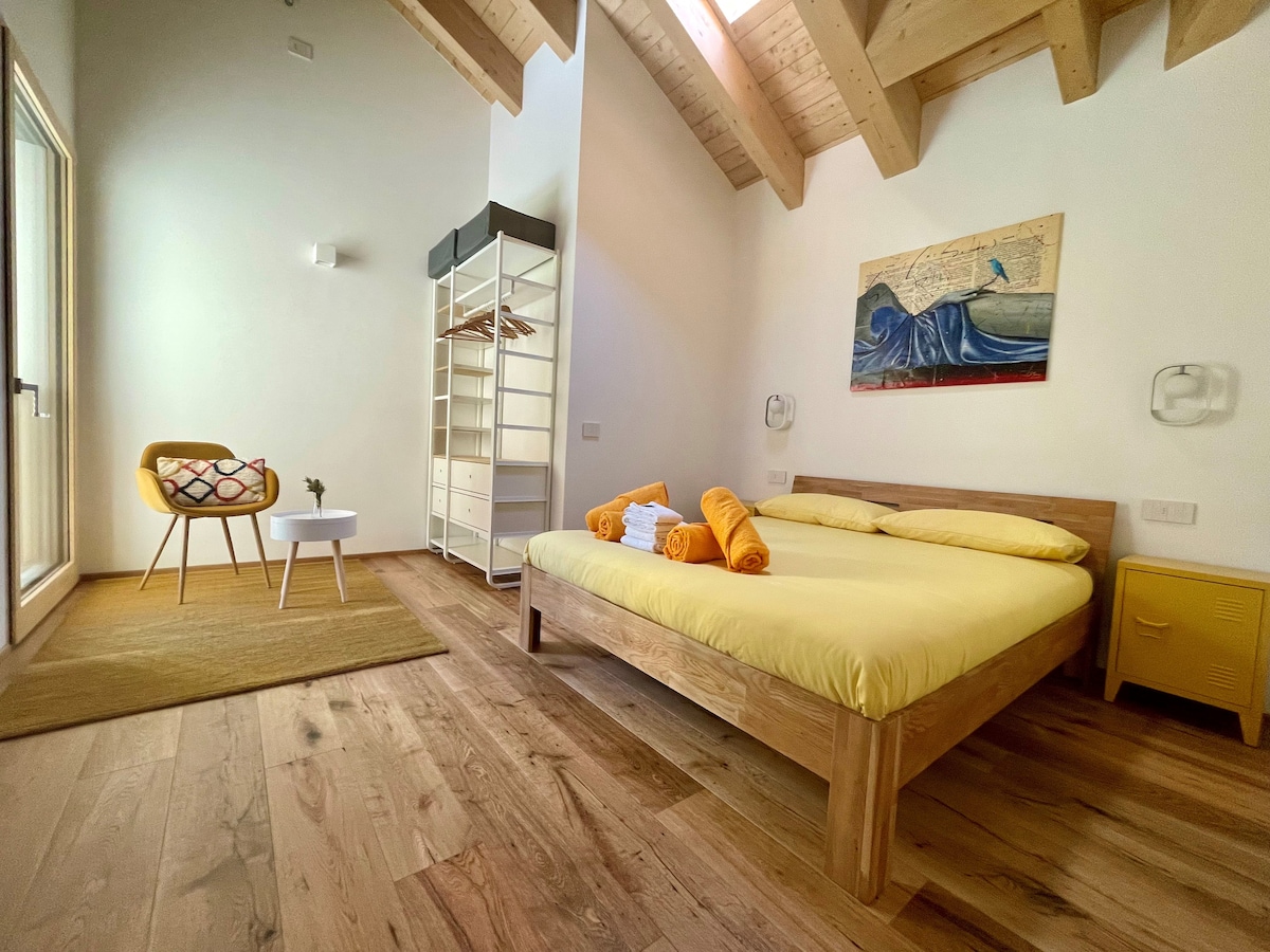 A spacious bedroom is highlighted with natural wooden flooring and a high ceiling with exposed beams. The room includes a yellow bed with neatly arranged towels, a bright armchair with a patterned cushion, and a small table, providing a warm and comfortable environment.