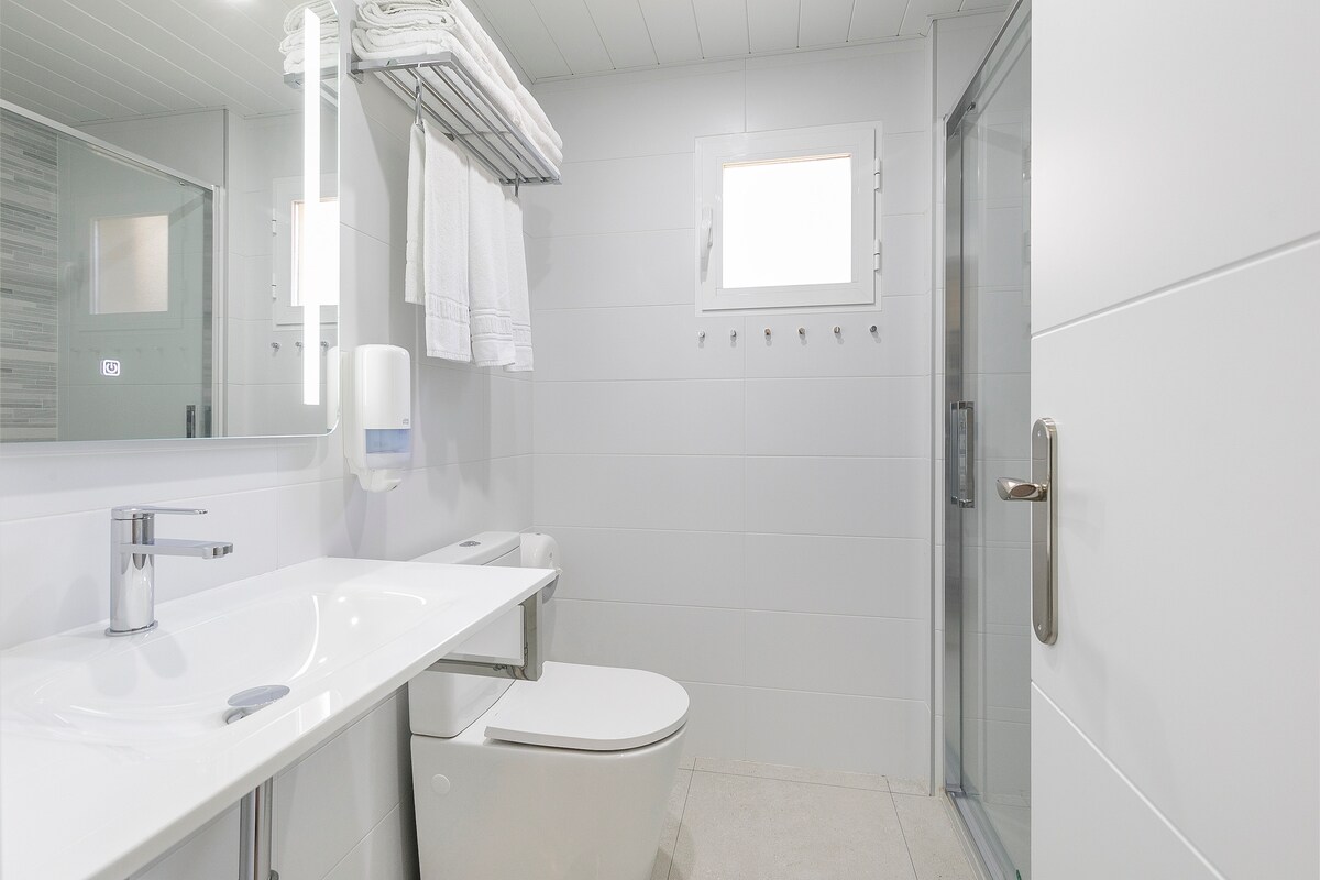 A modern bathroom features a sleek white sink with a polished chrome faucet. Soft white towels hang neatly on a rod above the sink. A glass shower enclosure is visible, along with a toilet positioned against the wall. Natural light enters through a small window.