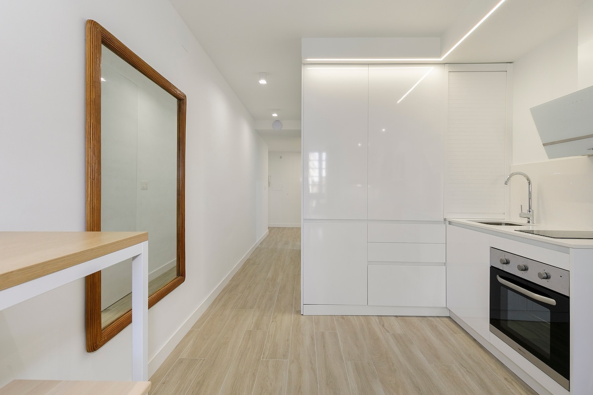 A modern kitchen area with sleek white cabinetry is showcased, featuring a blend of appliances including an oven and a stove. A full-length mirror adorns the adjacent wall, reflecting the light from the bright, neutral-toned interior. The flooring is a light wood finish, adding warmth to the space.