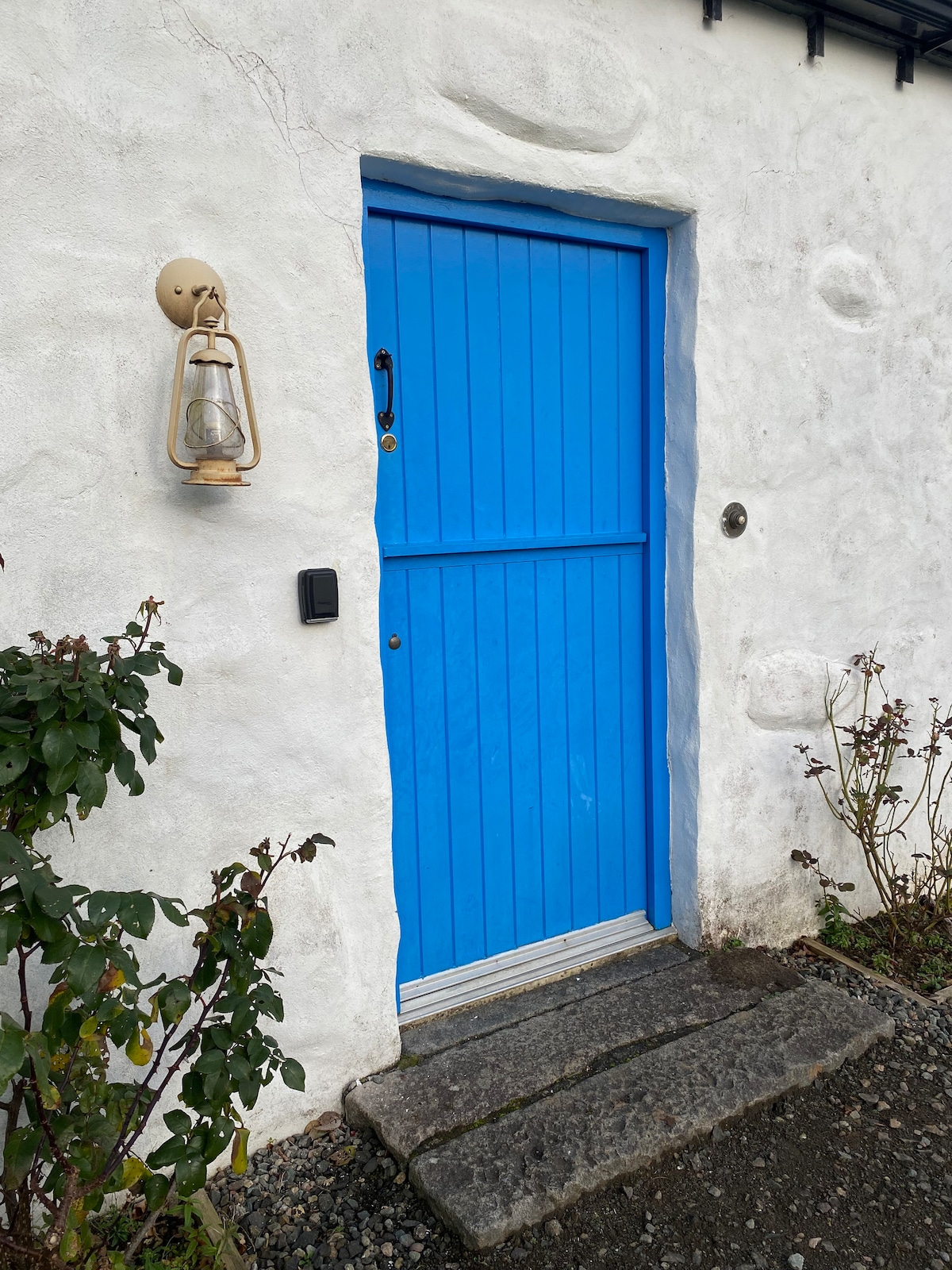 A traditional cottage entrance is highlighted by a vibrant blue door, framed by a white plastered exterior. A lantern is mounted beside the door, and a stone pathway leads to the entrance, surrounded by greenery and quaint stone details.