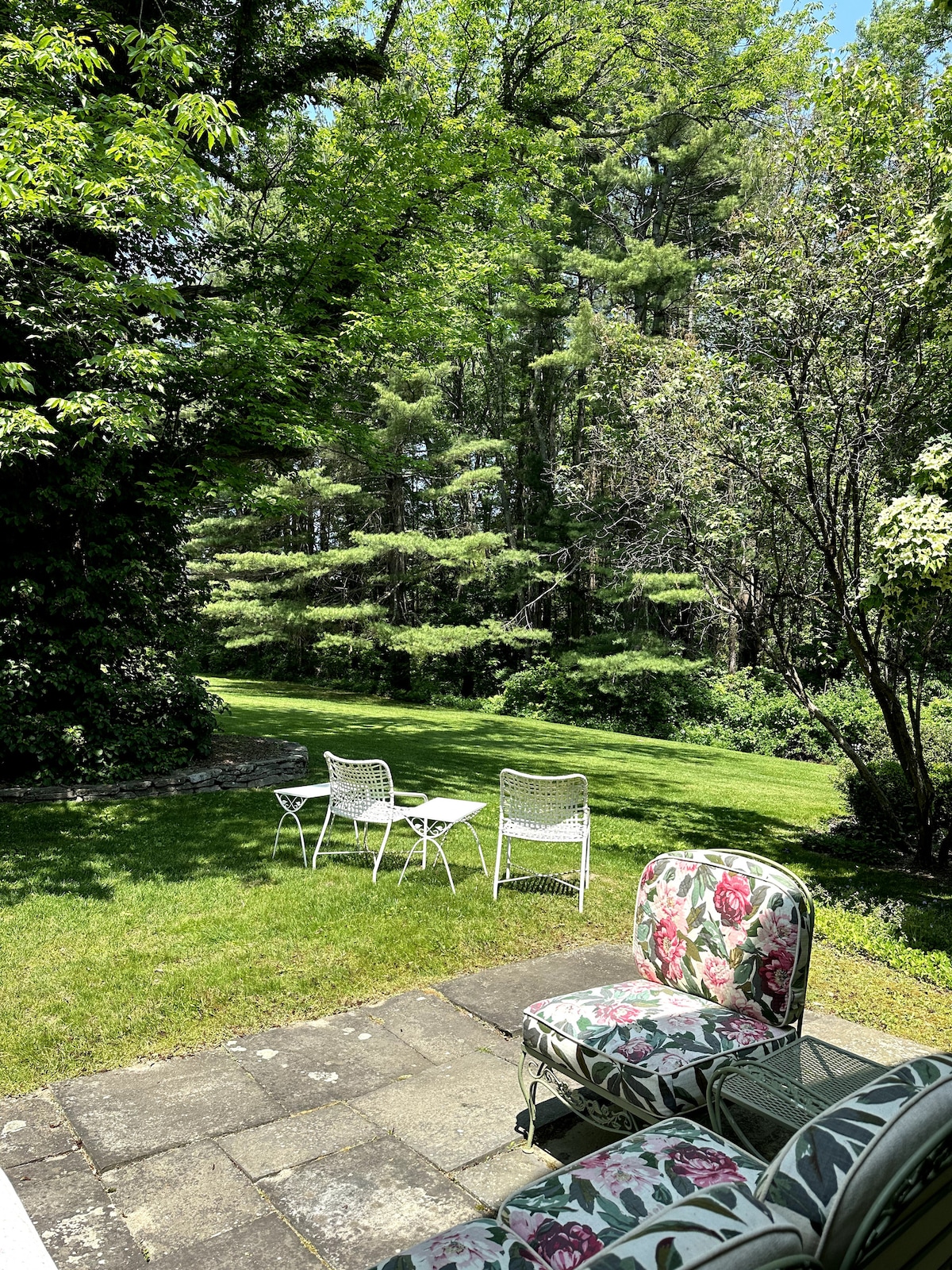 A tranquil outdoor seating area is presented, featuring floral-patterned chairs and a small table on a stone patio. Lush green grass extends into a wooded area beyond, where various trees provide shade and a serene backdrop.