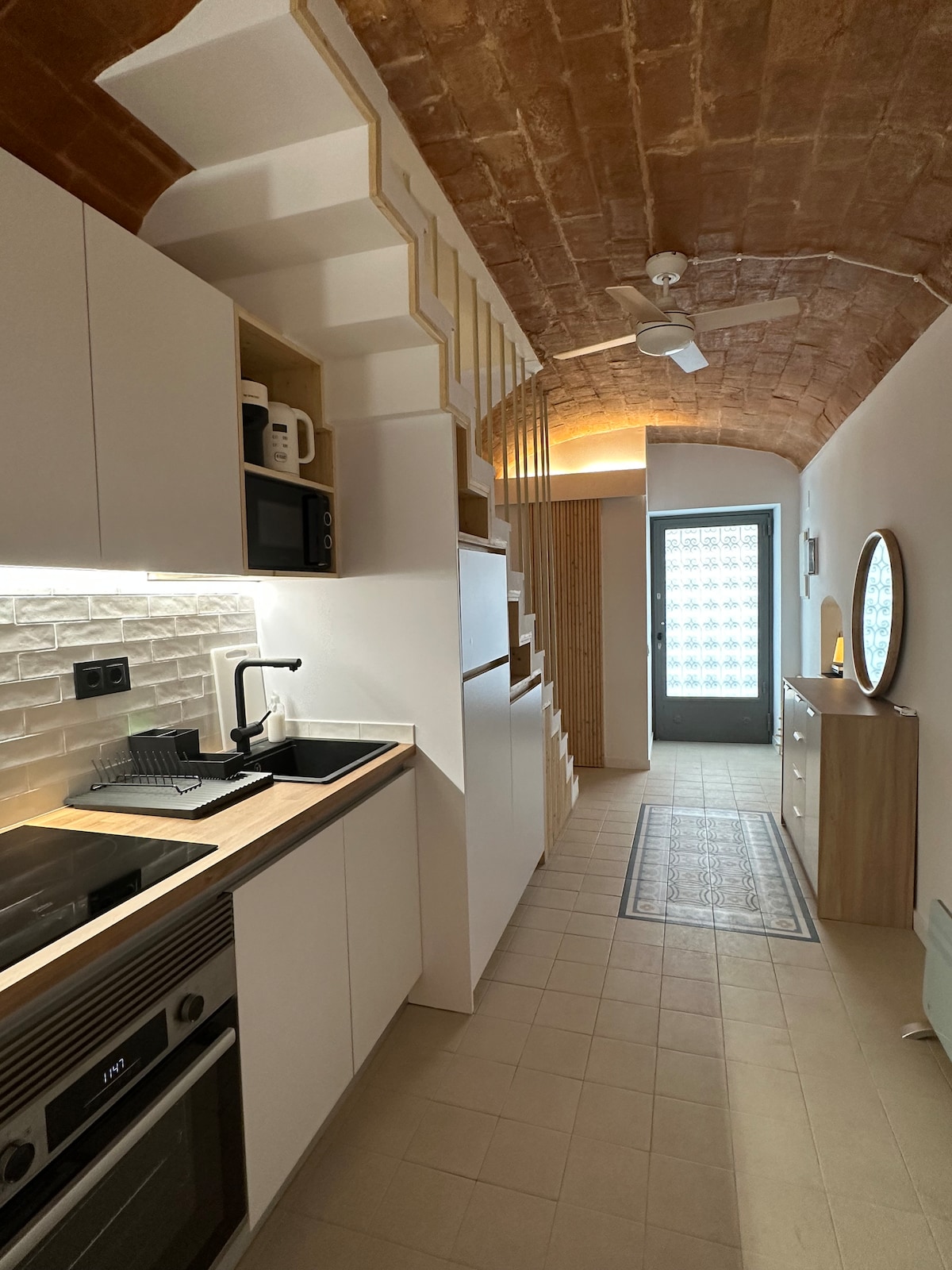 A modern kitchen features sleek white cabinetry and a wooden countertop. Under-cabinet lighting highlights the textured brick wall. A ceiling fan is visible above, and a doorway leads to a space with patterned glass, while a round mirror reflects the room's design.