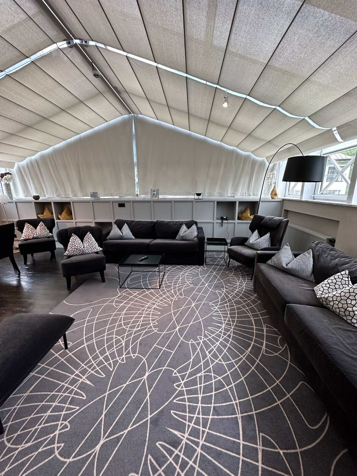 The airy living area features a collection of modern sofas adorned with patterned cushions. A sleek coffee table is positioned on a stylish area rug, while large windows are covered by soft, white drapes, allowing natural light to fill the space.