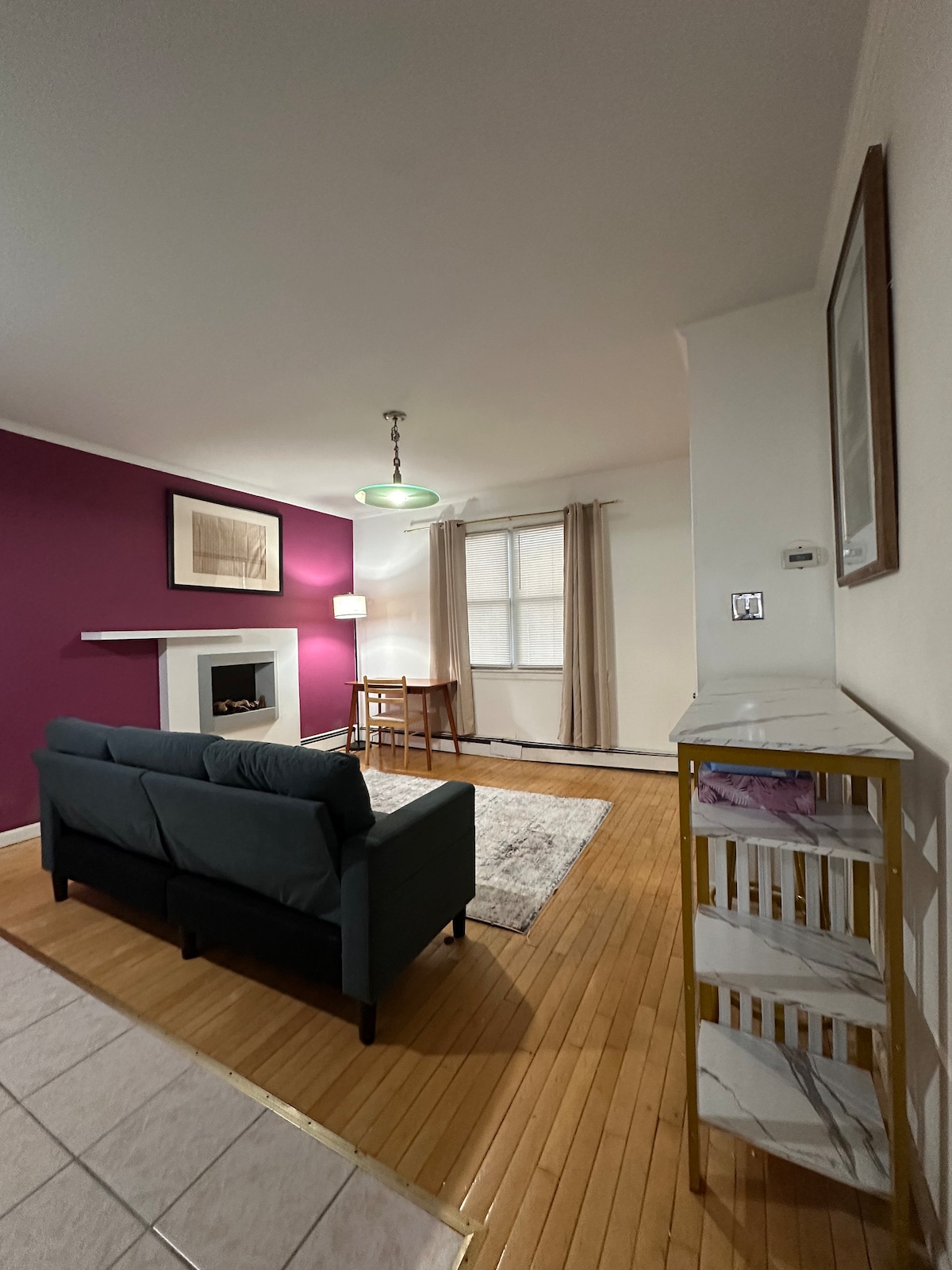 A modern living space features a comfortable black sofa facing a decorative fireplace against a deep purple wall. A small dining table with chairs is visible under a pendant light, while a window allows natural light to fill the area. A shelf adds storage and organization.