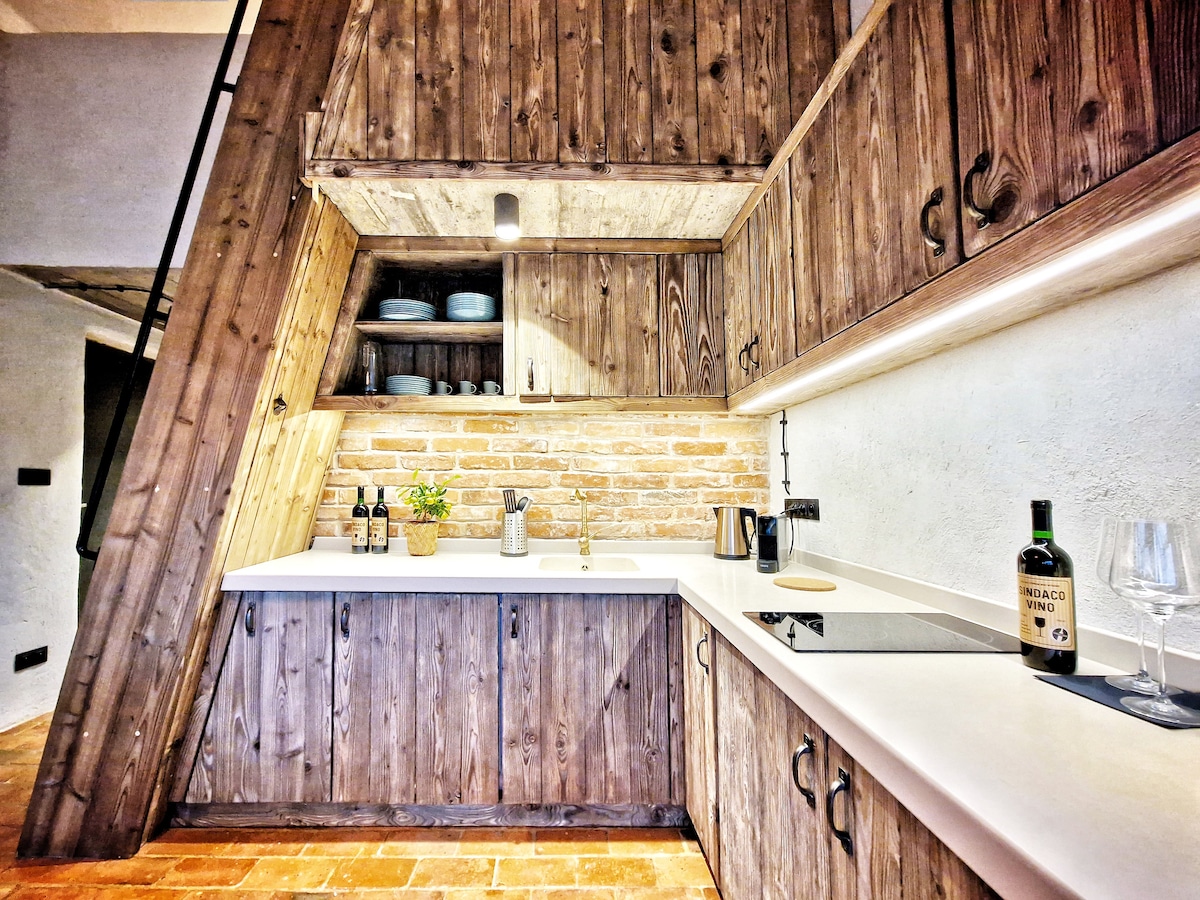 A modern kitchen features natural wooden cabinetry and a white countertop. Open shelving displays dishware and plants. A sleek stove is integrated into the counter. Stone walls provide a rustic backdrop, while soft lighting enhances the functional space.