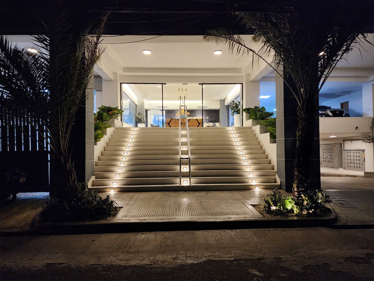 An inviting entrance is showcased, featuring wide steps illuminated by soft lighting. Lush greenery lines the sides, creating a welcoming path. Large glass doors are visible, hinting at a spacious interior. The overall design conveys a modern and accessible atmosphere.