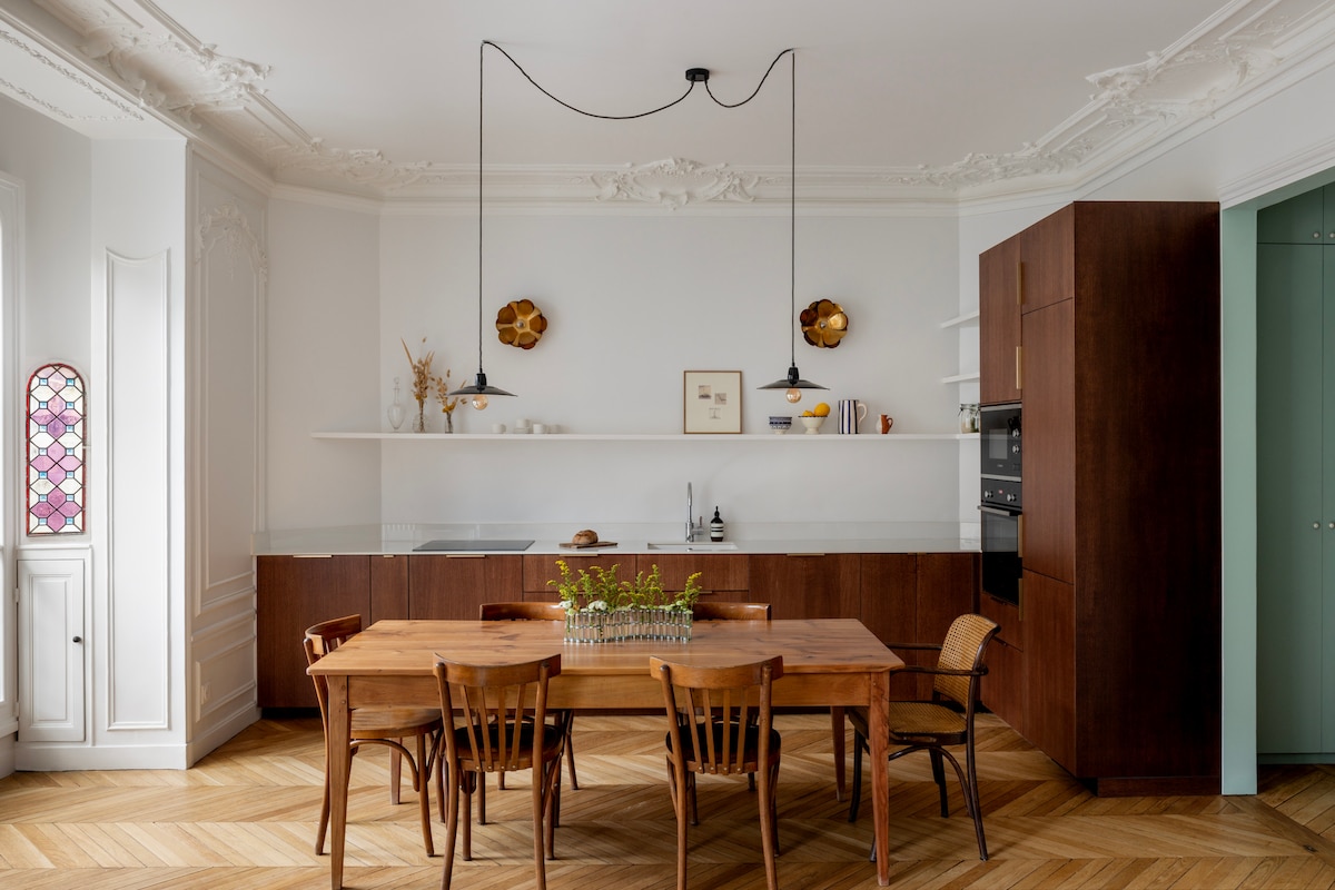 A spacious kitchen and dining area features a large wooden dining table surrounded by several chairs. The walls are accented with ornate details, and modern lighting hangs from the ceiling. A minimalist kitchen counter is equipped with essential appliances, and decorative elements are displayed on open shelving.