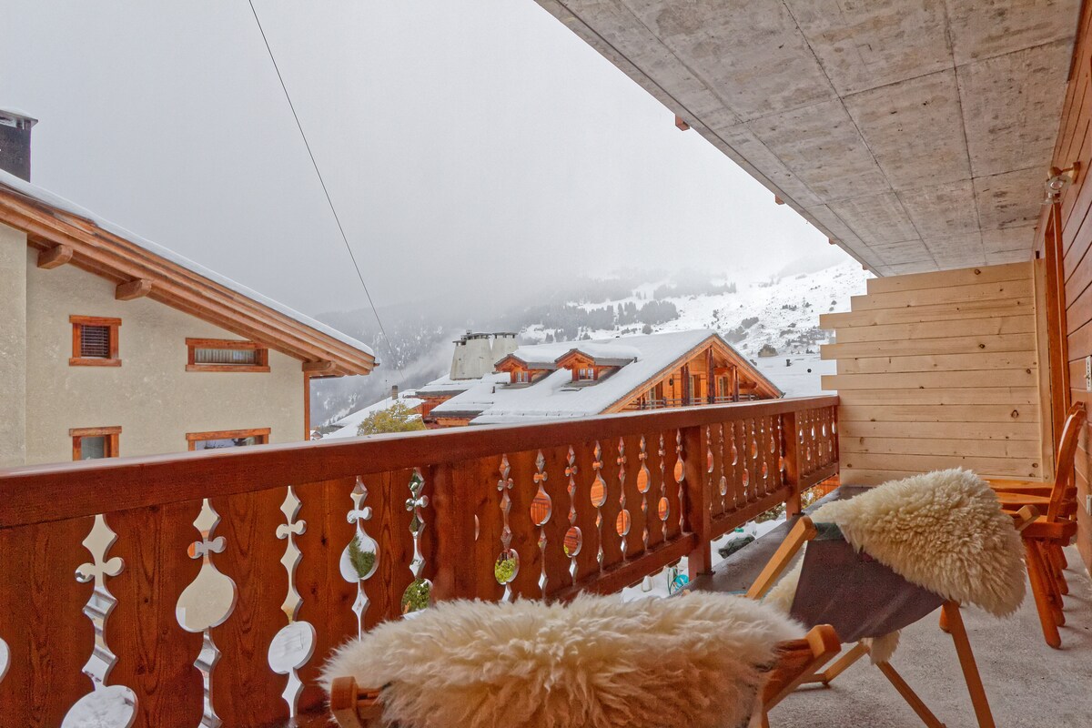 A spacious balcony features wooden railings with traditional carvings, offering views of the snow-covered landscape and nearby chalets. Two comfortable chairs are positioned on the balcony, accompanied by soft, textured blankets. The muted light of an overcast sky creates a quiet and serene ambiance.