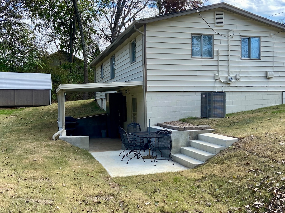An outdoor covered patio is seen adjacent to the home, featuring a small dining set with four chairs and a table. Steps lead down from the patio to the grassy yard, which is bordered by trees. A barbecue grill is visible in the corner.