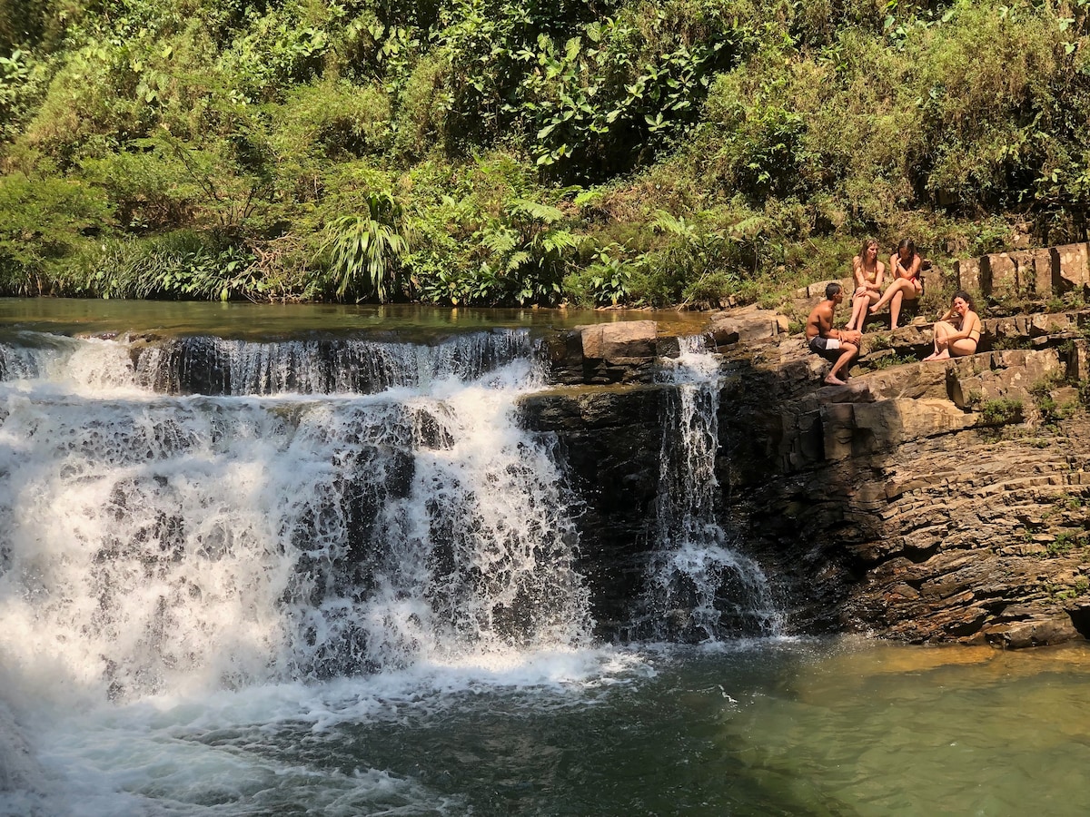 A group of individuals is seated on the rocks beside a flowing waterfall, surrounded by lush greenery. The water cascades over a series of natural stone ledges, creating a serene atmosphere in this tranquil outdoor setting.