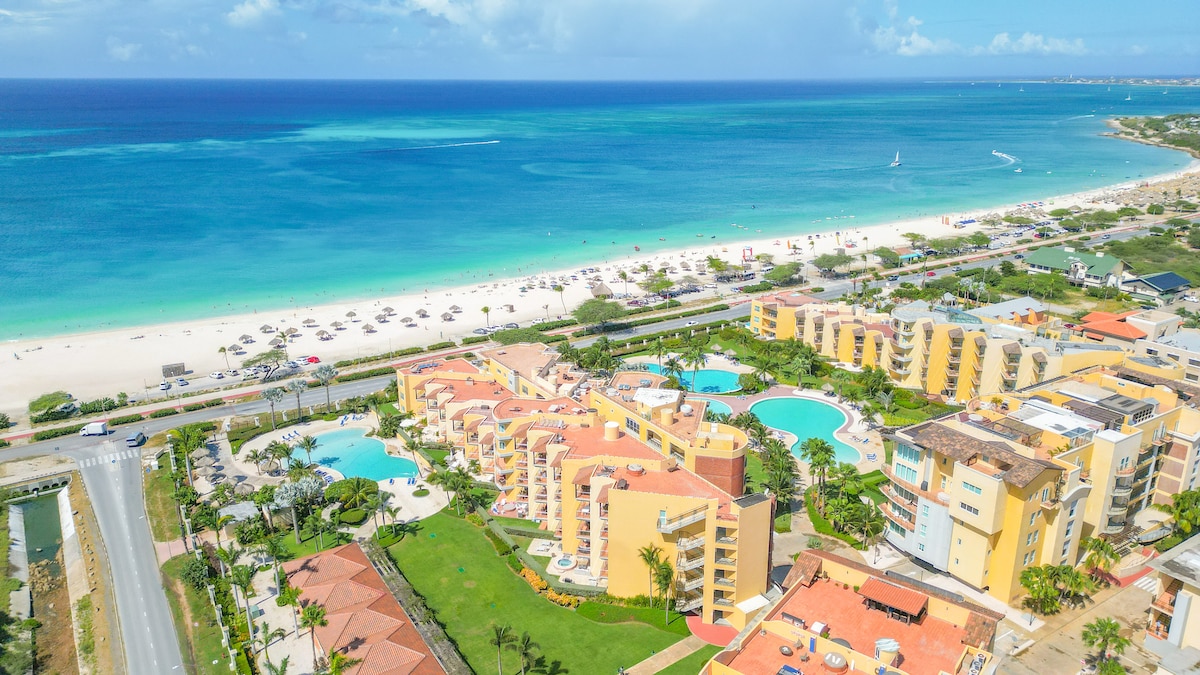 An aerial view showcases the Oceania Residences, situated directly on Eagle Beach. The expansive white sand beach extends alongside clear blue waters. The resort features inviting pools surrounded by palm trees, highlighting the tranquil atmosphere and seamless integration with the beach landscape.
