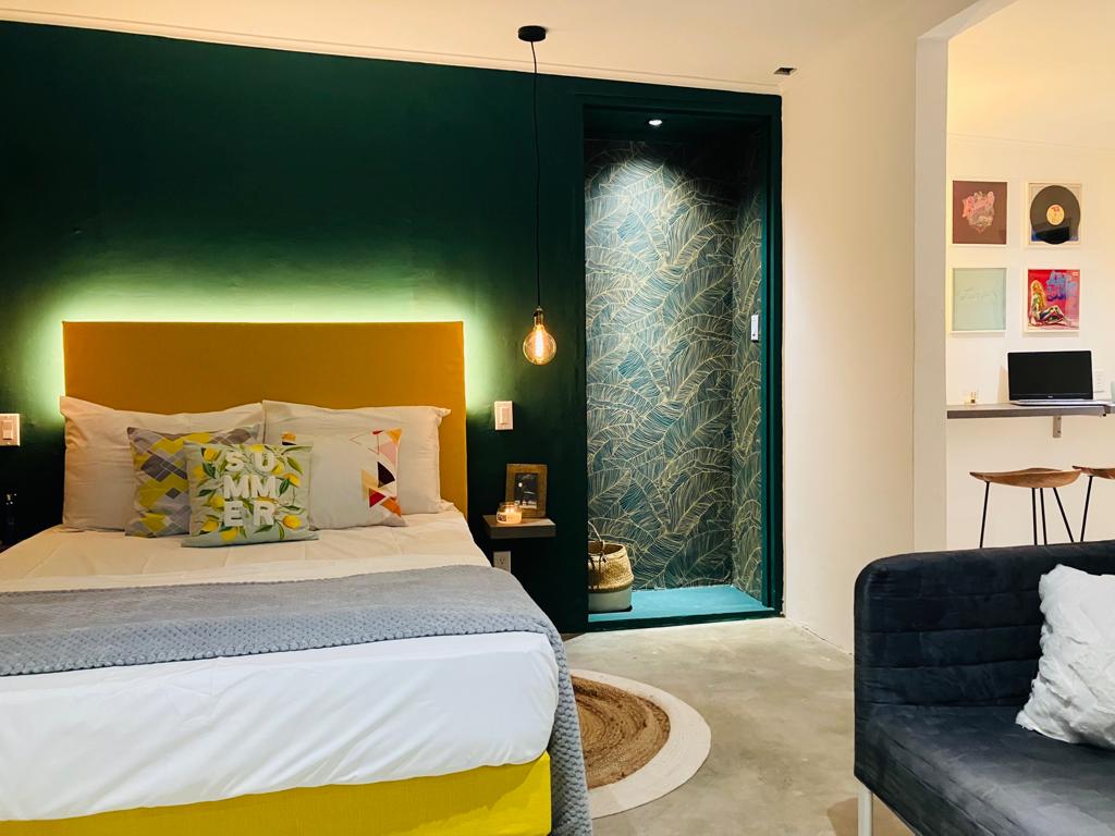 A cozy bedroom features a double bed adorned with decorative throw pillows and a modern bedside table. A wall in dark green provides a bold backdrop, while warm lighting enhances the space's comfort. A stylish couch complements the decor, and a doorway leads to the bathroom.