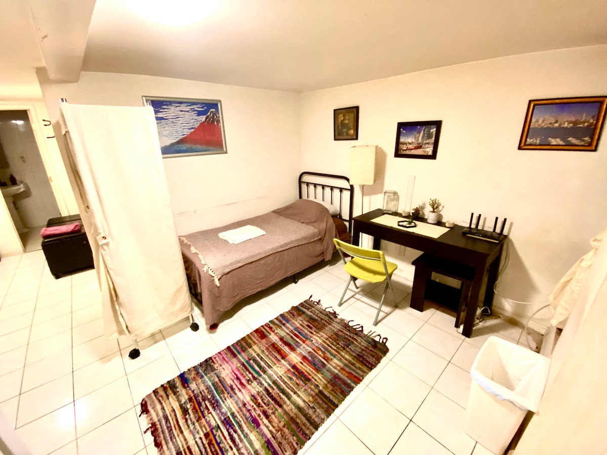 A small, private room is equipped with a single bed and a desk with a chair. A colorful area rug is placed on the tile floor, and artwork adorns the walls. A partition provides privacy, while a waste bin sits unobtrusively nearby.
