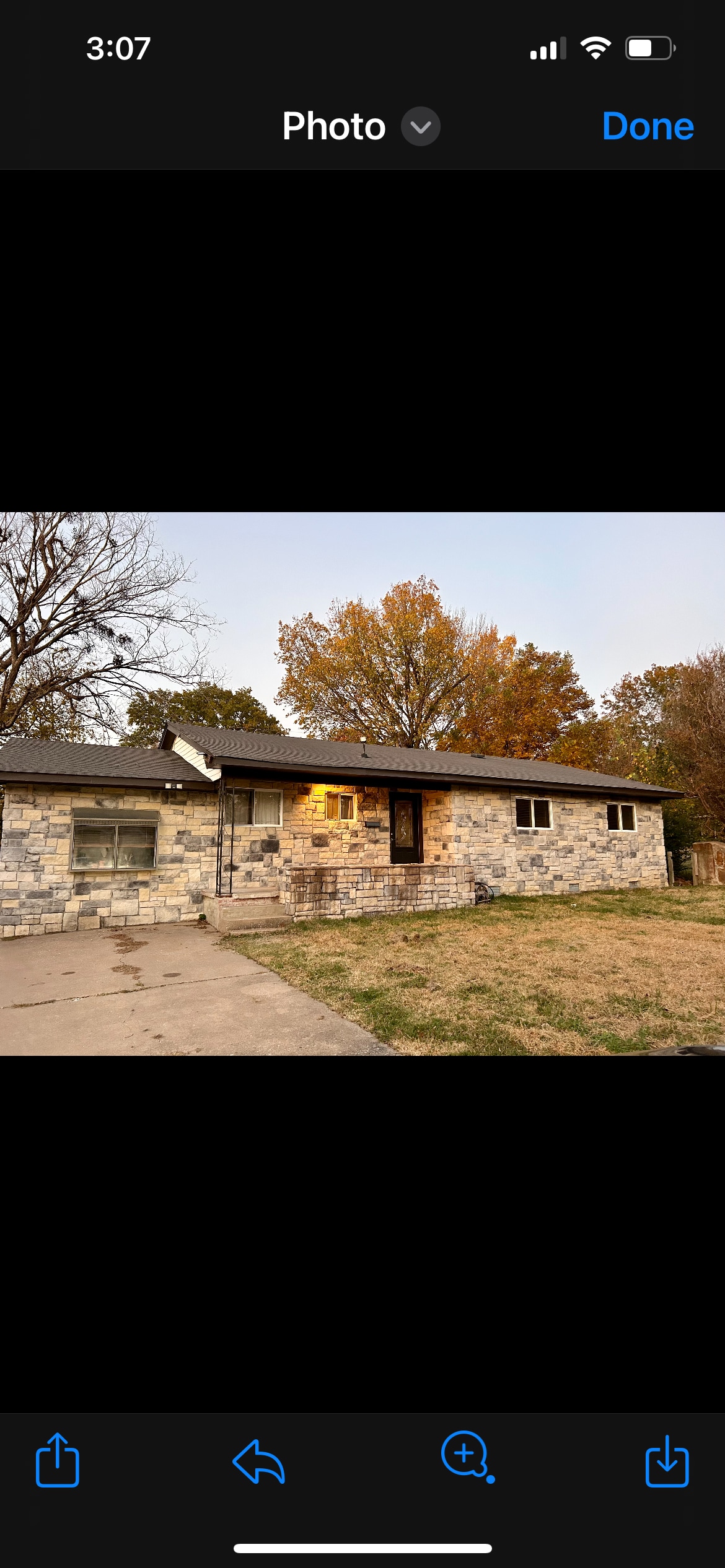 The exterior of a single-story stone house showcases large windows and a welcoming front door under a soft evening sky. Mature trees with autumn foliage frame the property, and a paved driveway leads to the entrance, providing a sense of approachability.