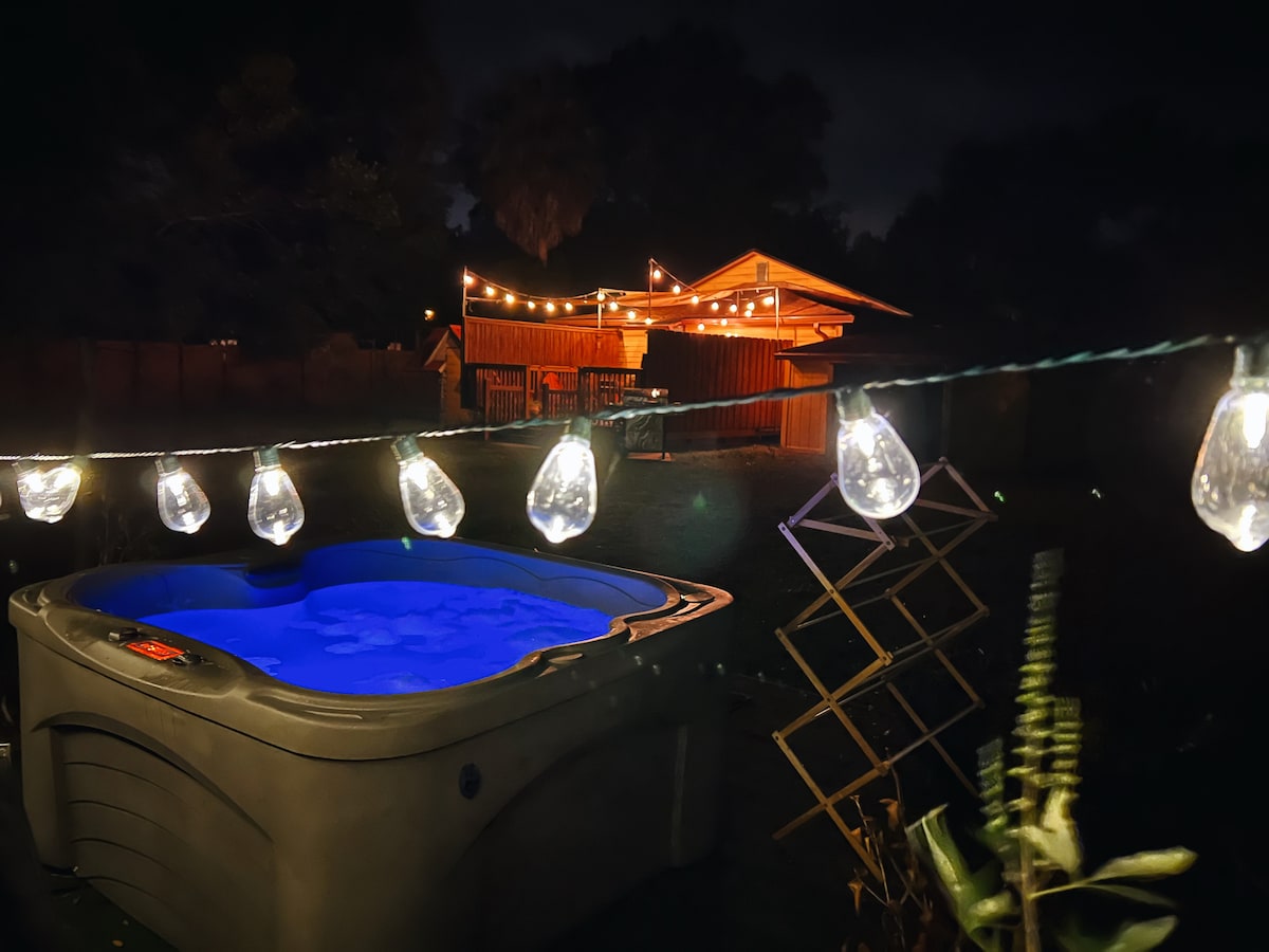 A hot tub glows in soft blue light under string lights, creating a serene outdoor space. A wooden structure with a patio is visible in the background, surrounded by greenery, providing an inviting atmosphere for relaxation.