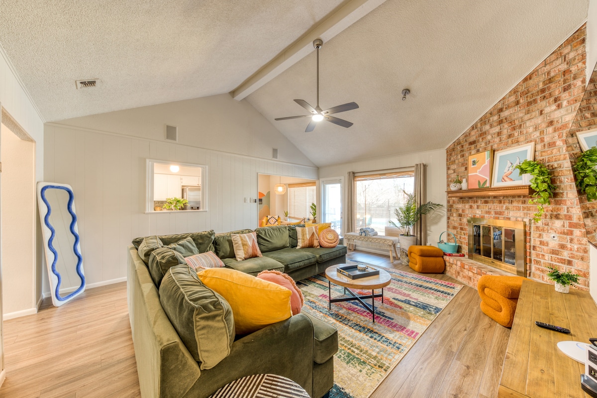 The living area features a spacious sectional sofa adorned with colorful throw pillows, set against a backdrop of a brick fireplace. Natural light fills the room through large windows, and a vibrant area rug adds warmth to the hardwood flooring.