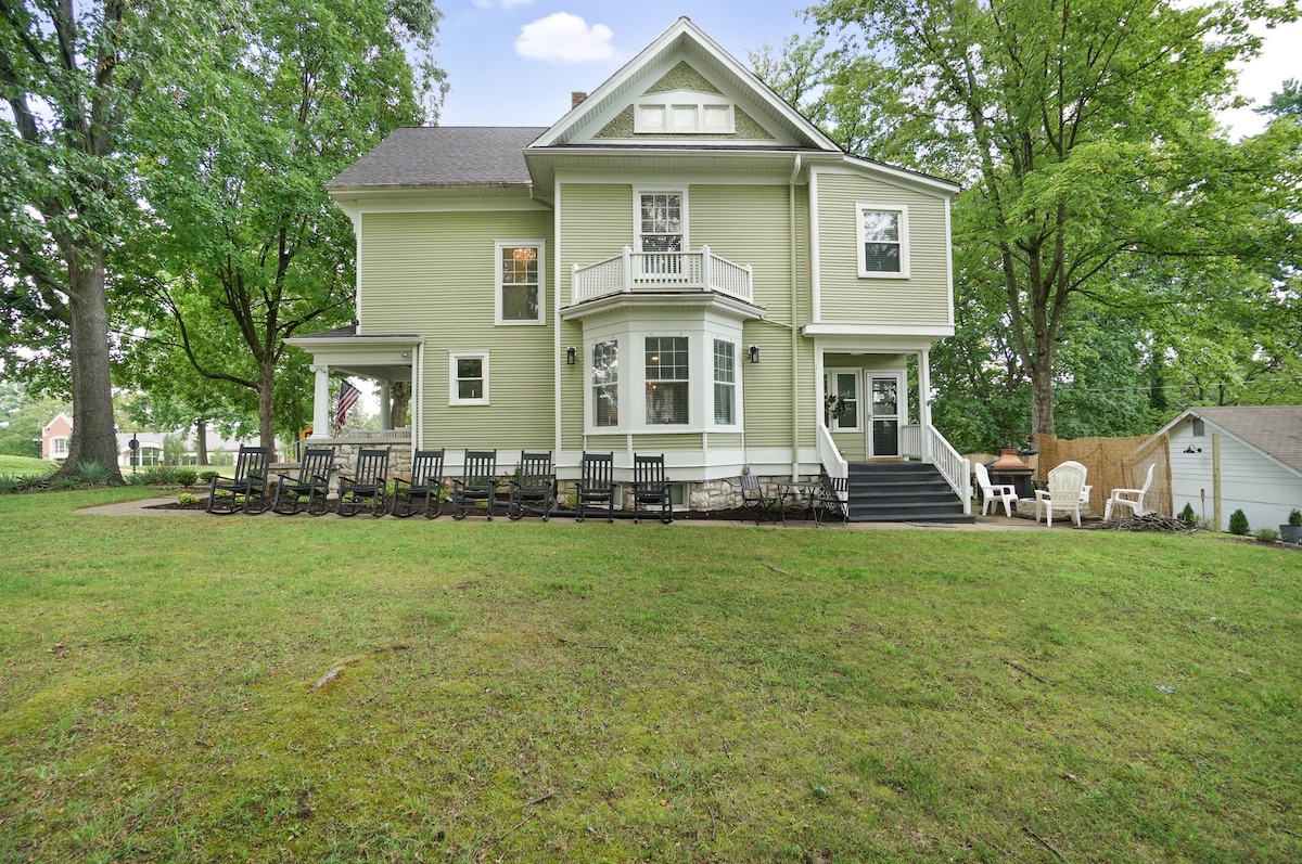 A spacious exterior view of a green house surrounded by lush greenery. The yard features multiple rocking chairs arranged neatly, inviting relaxation. The two-story structure showcases a balcony on the second floor, with large windows that provide ample natural light.