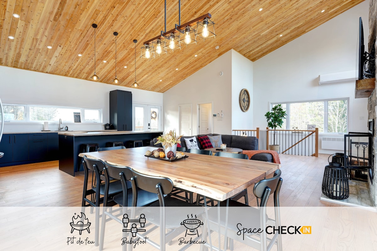 The spacious dining area features a large wooden table with seated chairs, surrounded by inviting natural light from expansive windows. A cozy living area, visible in the background, showcases a modular sofa and a unique ceiling design with wood paneling.