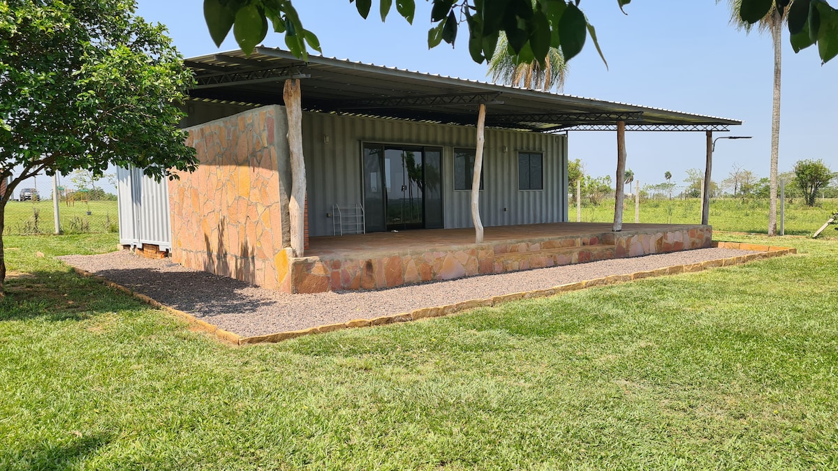 A modern structure constructed from stone and metal is set among greenery. The spacious outdoor area features a wide wooden deck, bordered by a gravel path. Large glass doors provide a view of the interior, framed by trees offering gentle shade.