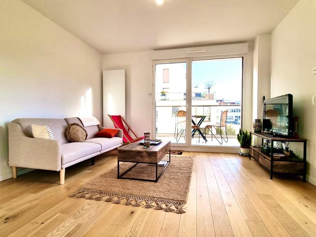 A modern living area features a comfortable sofa and a coffee table on a woven rug. Large glass doors open to a balcony, allowing ample natural light. Minimalist decor is observed, with a television stand and greenery enhancing the space.
