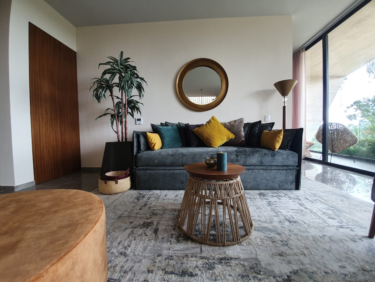 A living area features a dark sofa adorned with colorful cushions, complemented by a round coffee table made of natural materials. A large mirror reflects the space, while a green plant adds a touch of nature. Floor-to-ceiling windows invite ample natural light and offer views of the outdoors.
