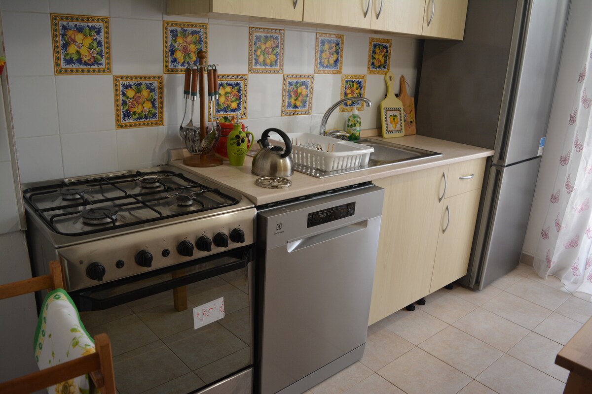 A functional kitchen setup is visible, featuring a gas stove, a dishwasher, and a stainless-steel sink. Colorful tiled backsplash adds character. Counter space is utilized for food preparation, with kitchenware neatly arranged. A window draped with light fabric allows natural light to enter the space.