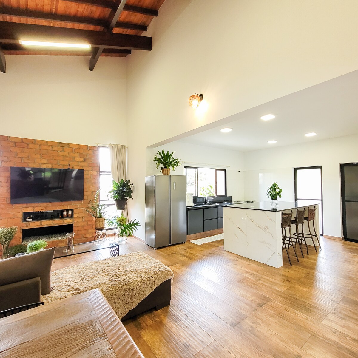 The open-concept living area features a combination of natural wood and modern elements. A cozy seating area with plants is complemented by a brick accent wall and a mounted television. The kitchen, equipped with sleek cabinetry and an island with seating, invites gatherings.