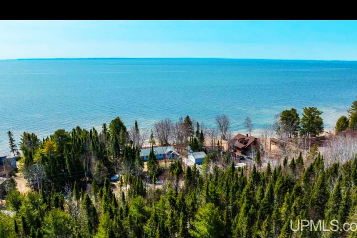 An aerial view showcases a serene lakeside setting with clear blue waters. Lush green trees surround quaint structures, blending harmoniously with the natural landscape. A sandy beach is visible along the shoreline, creating a tranquil retreat atmosphere.