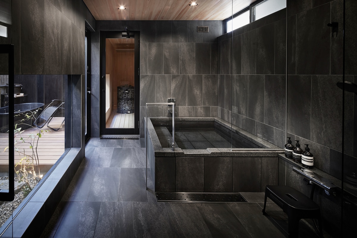 A serene indoor bathing area features dark stone walls and flooring, complemented by a square bathtub filled with water. Natural light filters in through large windows, illuminating the space. A doorway leads to an outdoor deck, enhancing the tranquil atmosphere.