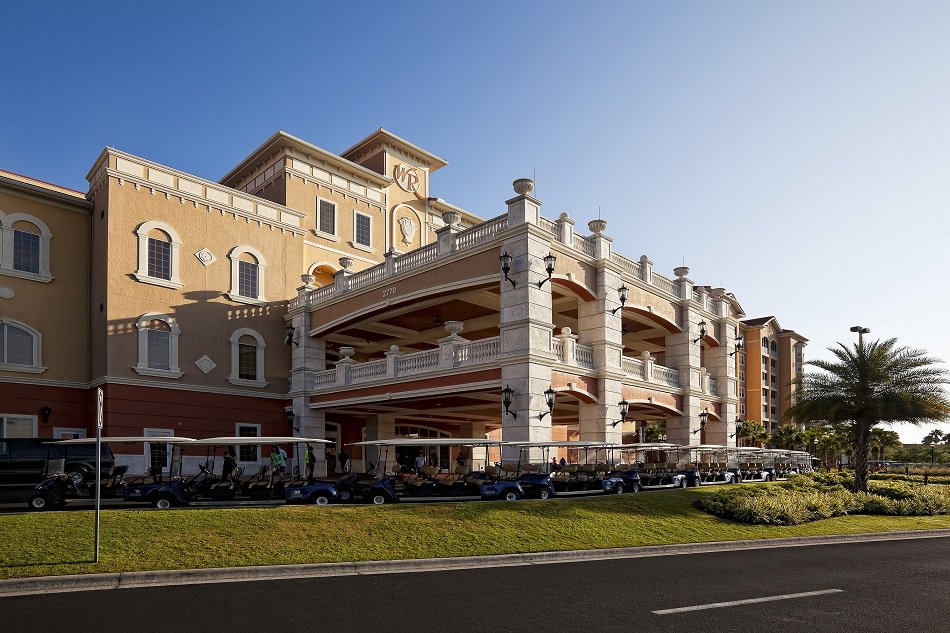 A grand resort building showcases elegant architecture with decorative columns and balconies. The structure features multiple levels, while rows of golf carts are neatly parked in front. Lush greenery, including palm trees, frames the scene under a clear blue sky.
